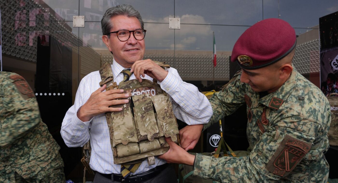 Ricardo Monreal ajusta un chaleco táctico con ayuda de un militar durante una exhibición.